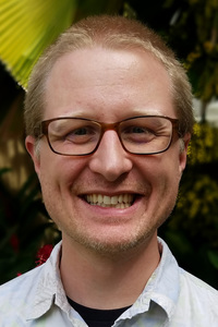 A smiling headshot of Joe Oppegaard, who has shaved blonde hair, brown frame glasses, a little bit of beard stubble, and a shirt with a collar.